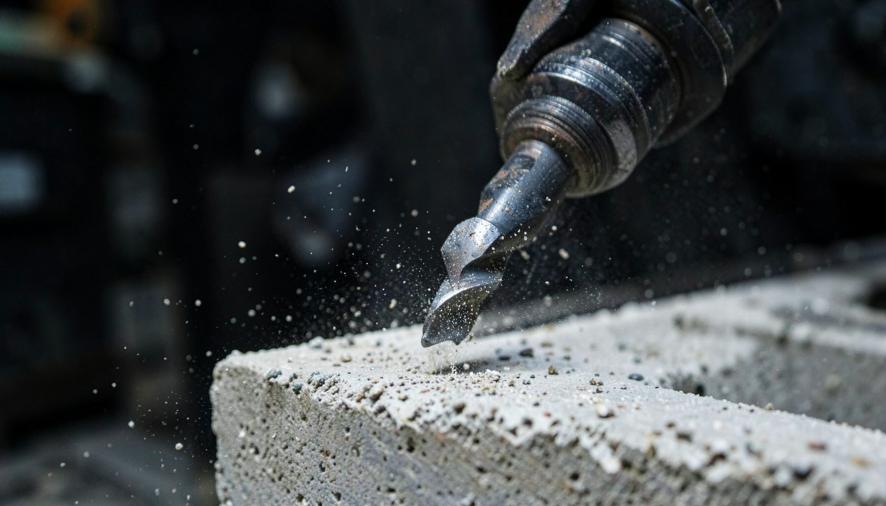 Close-up of a professional drill bit cutting into concrete with fine dust particles against a dark workshop backdrop